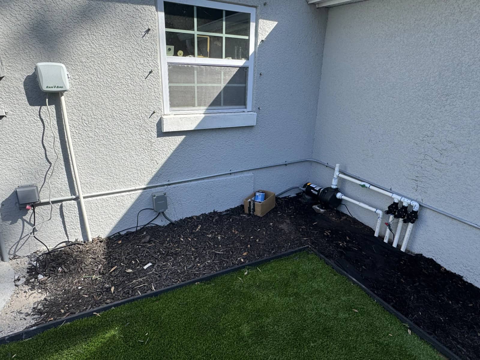 Irrigation equipment installed along a home exterior, including a Rain Bird controller, pumps, and PVC piping.